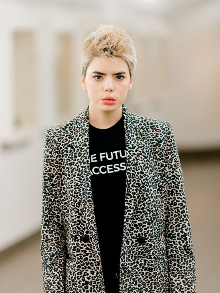 A photograph of Kristina McMullin taken by Michelle Peek of Bodies in Translation. She looks into the camera with a neutral, albeit a bit tired, look on her face. She has short dyed blond hair and dark brown eyebrows and eyes which contrast against her fair skin. She wears a black and white leopard blazer over a black shirt with white all caps lettering. Some of the lettering is obstructed, but the slogan reads ‘The Future Is Accessible’. Kristina stands apart from the out of focus background.
