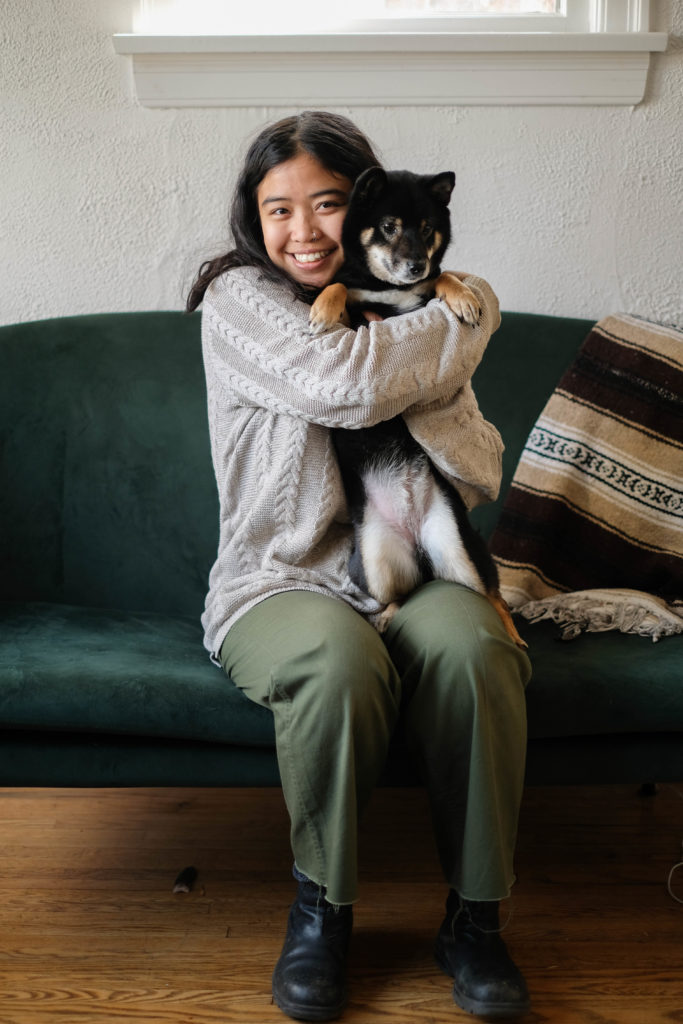 Image Description: A photograph of Alexis hugging their black and tan shiba inu, Satomi, sitting on an emerald green velvet couch. A striped woven blanket is draped along the back of the couch. The image captures Alexis’ full sitting body. Satomi is on her hind legs, balancing on one of Alexis’ legs, and Satomi’s underbelly is showing. Alexis is smiling at the camera with their face squished up against Satomi’s face. Satomi’s tan paws resting on Alexis’ wrapped arms. Alexis has wavy, mid-length brown hair that collects messily along her shrugged shoulders. Their skin is an almond tone. Alexis is wearing a knitted light beige sweater, green pants, and black boots.
