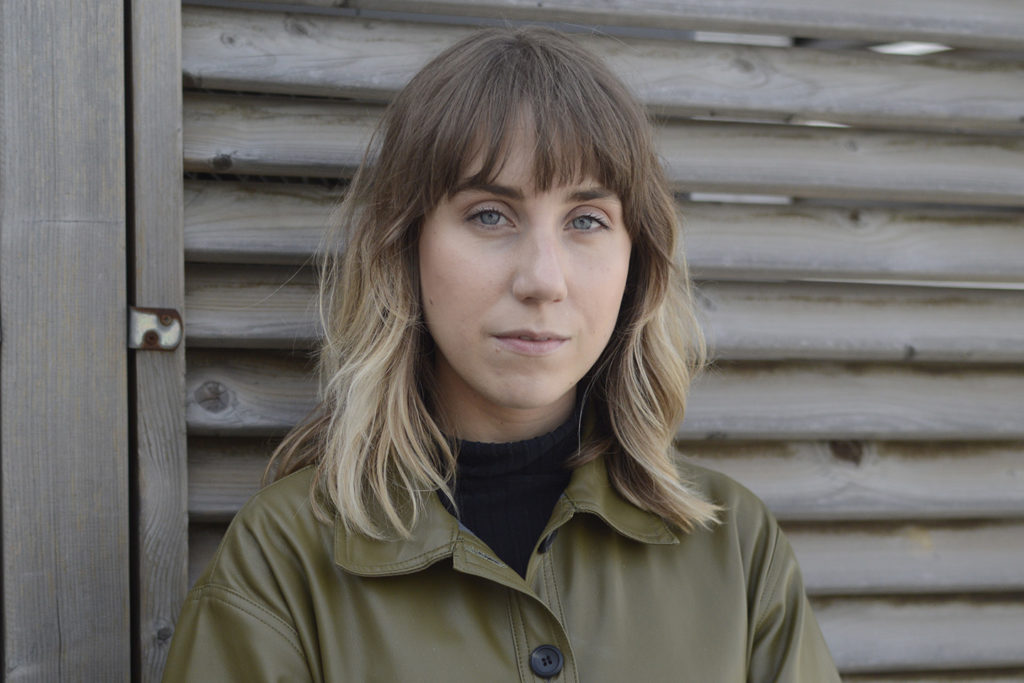 A Photo of Phillipa. She is a white woman with light brunette hair and blue eyes. She wears a green buttoned jacket and a black turtle neck top. She looks at the camera with a serious gaze while in front of a wood panelled wall. 